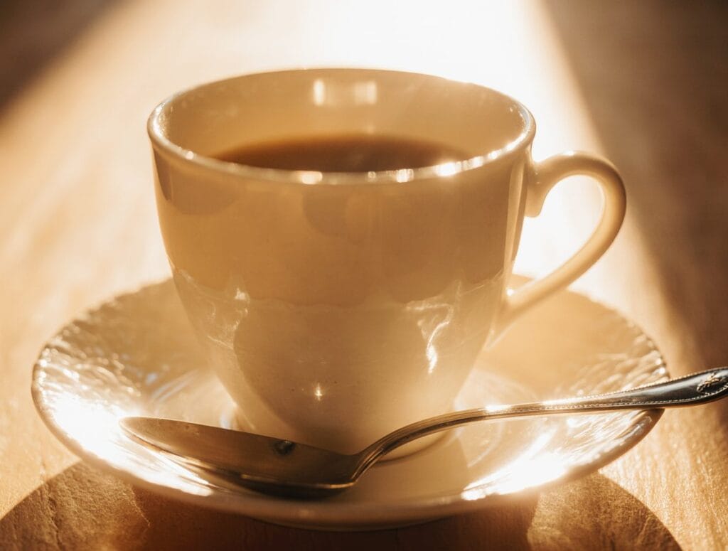 Taza de café blanca con cucharilla de metal en el plato, iluminada con luz dorada de atardecer. Edición generada con Krea 1.